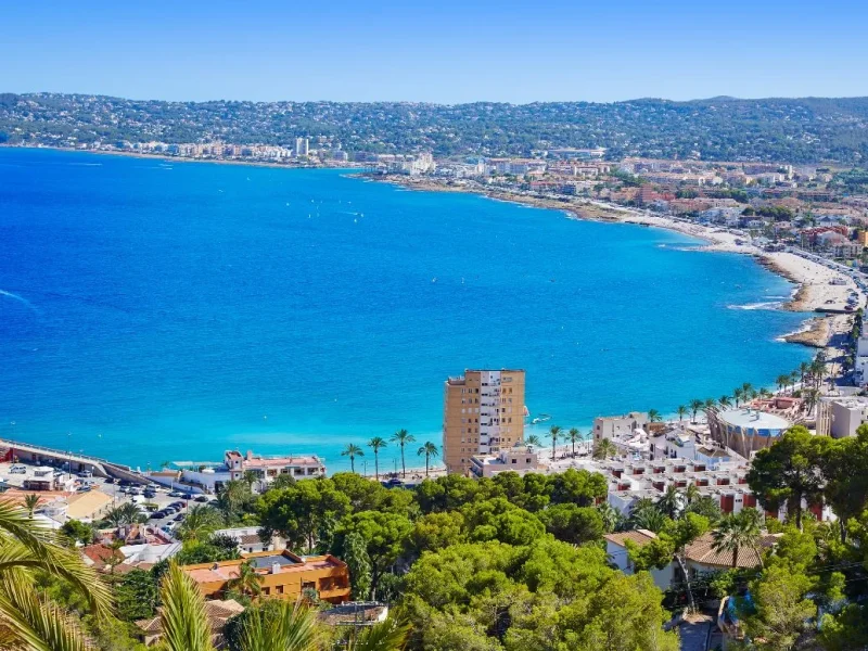 Vista panorámica de la costa de Jávea, con su playa, edificios y vegetación exuberante.