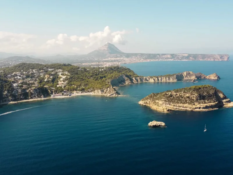 Panorámica aérea de la costa de Jávea, con colinas verdes salpicadas de residencias, una playa de arena y una isla rocosa destacada en el mar azul. Un velero y la estela de una lancha son visibles, con el Montgó al fondo. Panorámica aérea de la costa de Jávea, con colinas verdes salpicadas de residencias, una playa de arena y una isla rocosa destacada en el mar azul. Un velero y la estela de una lancha son visibles, con el Montgó al fondo.