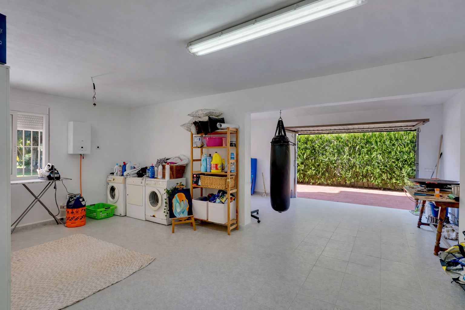 Amplio espacio multifuncional con zona de lavandería, área de gimnasio con saco de boxeo y acceso directo al exterior a través de una gran puerta de garaje.