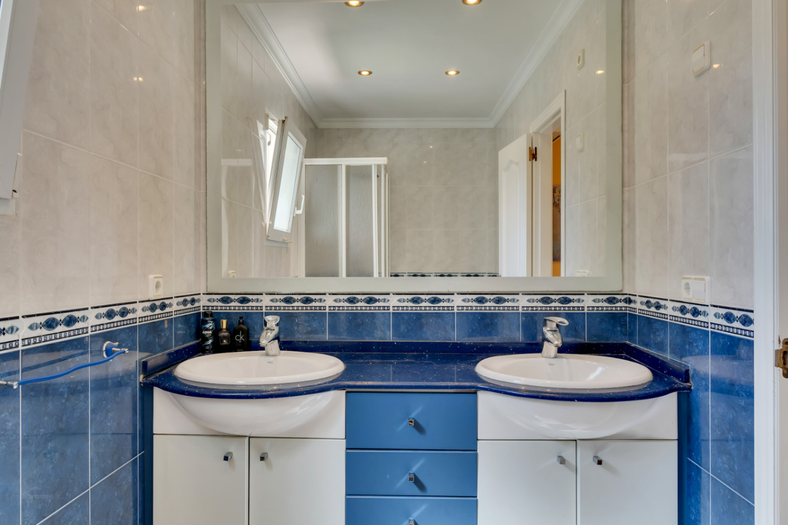 Baño con lavabo doble integrado en encimera azul, grifería cromada y mueble de almacenaje. Paredes alicatadas en tonos azules y blancos con espejo de gran formato y focos empotrados.