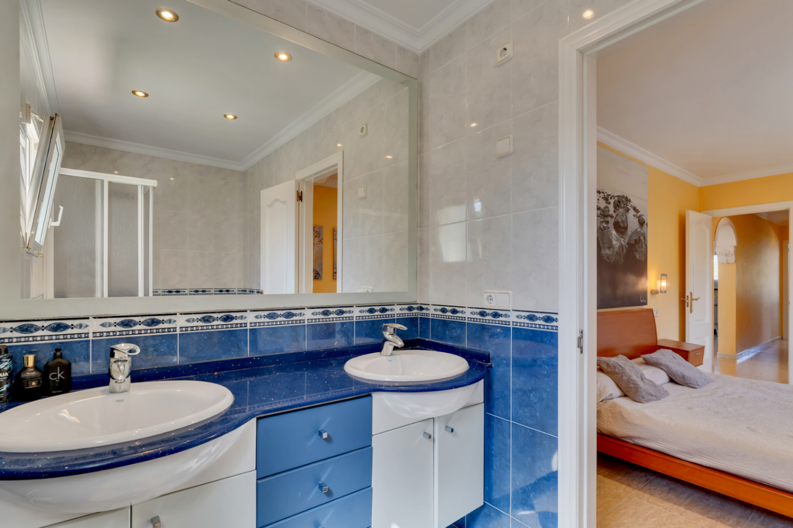 Baño en suite con lavabo doble, encimera azul y azulejos a juego. Incluye un gran espejo con focos empotrados y acceso directo al dormitorio principal.