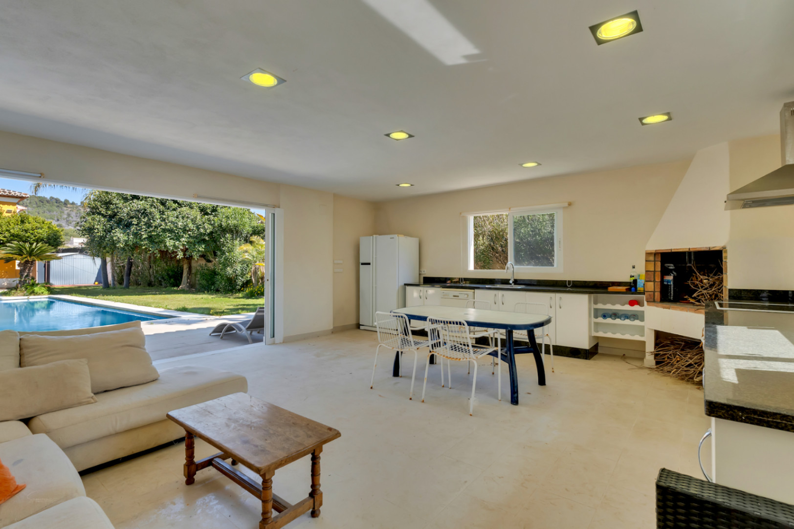 Cocina de verano integrada con zona de estar, equipada con barbacoa de obra y encimera de granito, abierta directamente a la terraza con piscina y jardín mediterráneo.