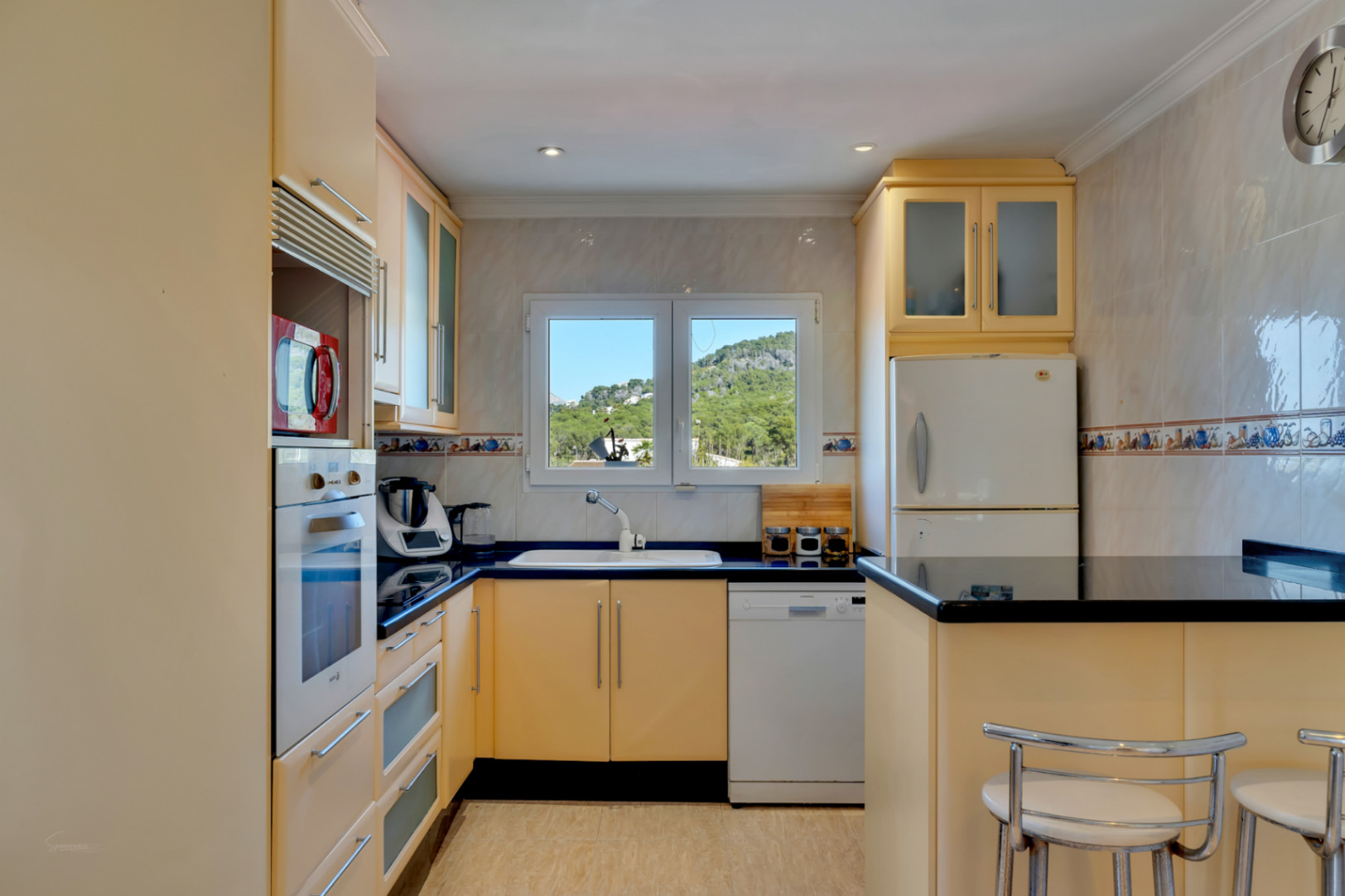 Cocina equipada con armarios en tono crema, encimeras de granito oscuro y una ventana central con vistas despejadas a las colinas de Jávea.