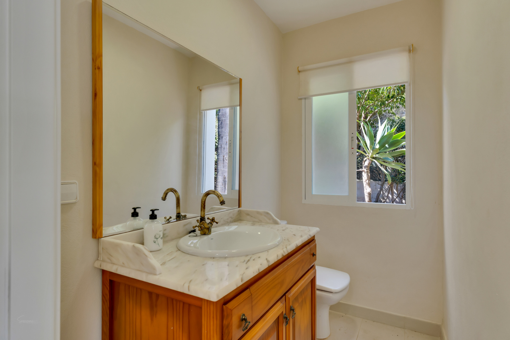 Cuarto de baño con mueble de madera, encimera de mármol veteado y grifería de estilo clásico. Una ventana exterior aporta luz natural y vistas a la vegetación del jardín.