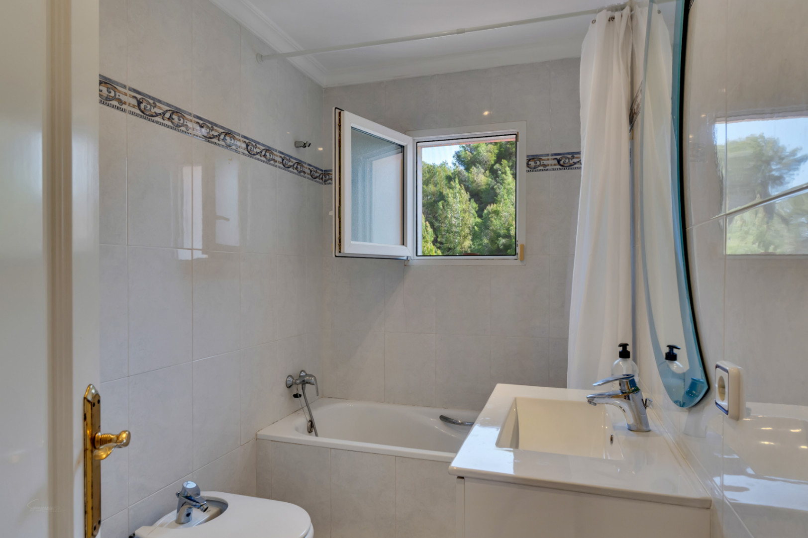 Cuarto de baño funcional con bañera, bidé y mueble de lavabo blanco. Incluye una ventana oscilobatiente con vistas a la vegetación y azulejos con cenefa decorativa.