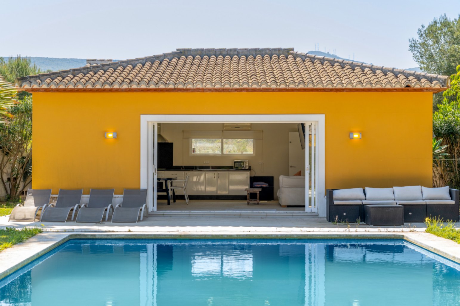 Edificio anexo junto a la piscina con cocina equipada, zona de estar y amplios ventanales correderos que integran el espacio con el solárium y las tumbonas.