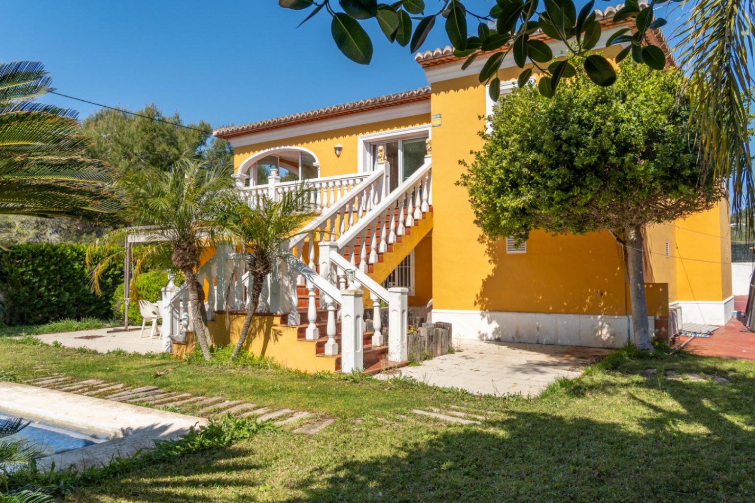 Fachada amarilla con escalera exterior de balaustrada blanca que conecta el jardín con la terraza superior. Entorno mediterráneo con palmeras y acceso directo a la piscina.