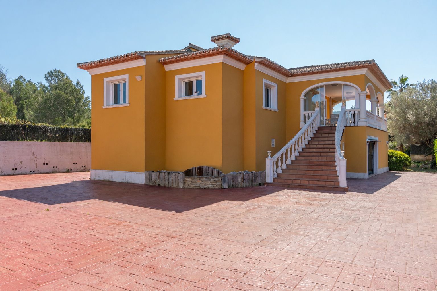 Fachada exterior de la villa en tono ocre con escalera de balaustrada blanca, naya arqueada y amplia zona de aparcamiento en hormigón impreso terracota.