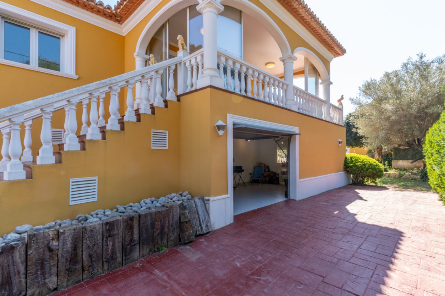 Fachada mediterránea con escalera exterior y balaustrada blanca que conduce a una naya arqueada sobre un garaje integrado y amplio patio pavimentado.