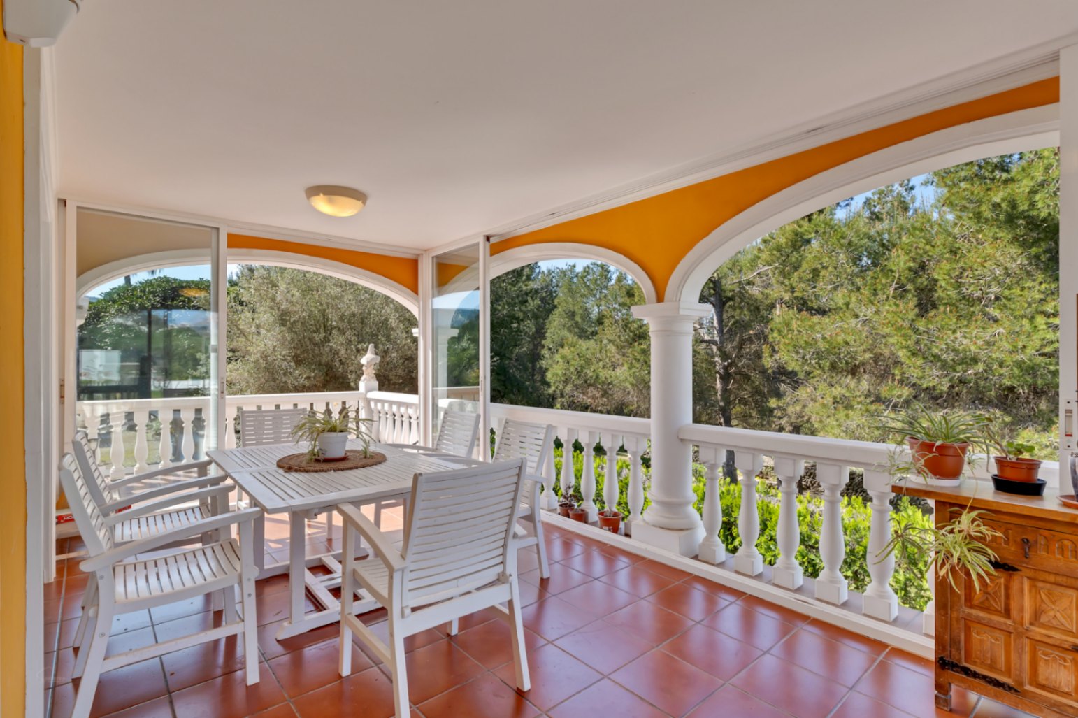 Terraza cubierta con arcos, suelos de terracota y cerramiento acristalado. Incluye comedor de madera blanca con vistas despejadas a la naturaleza y los pinos.