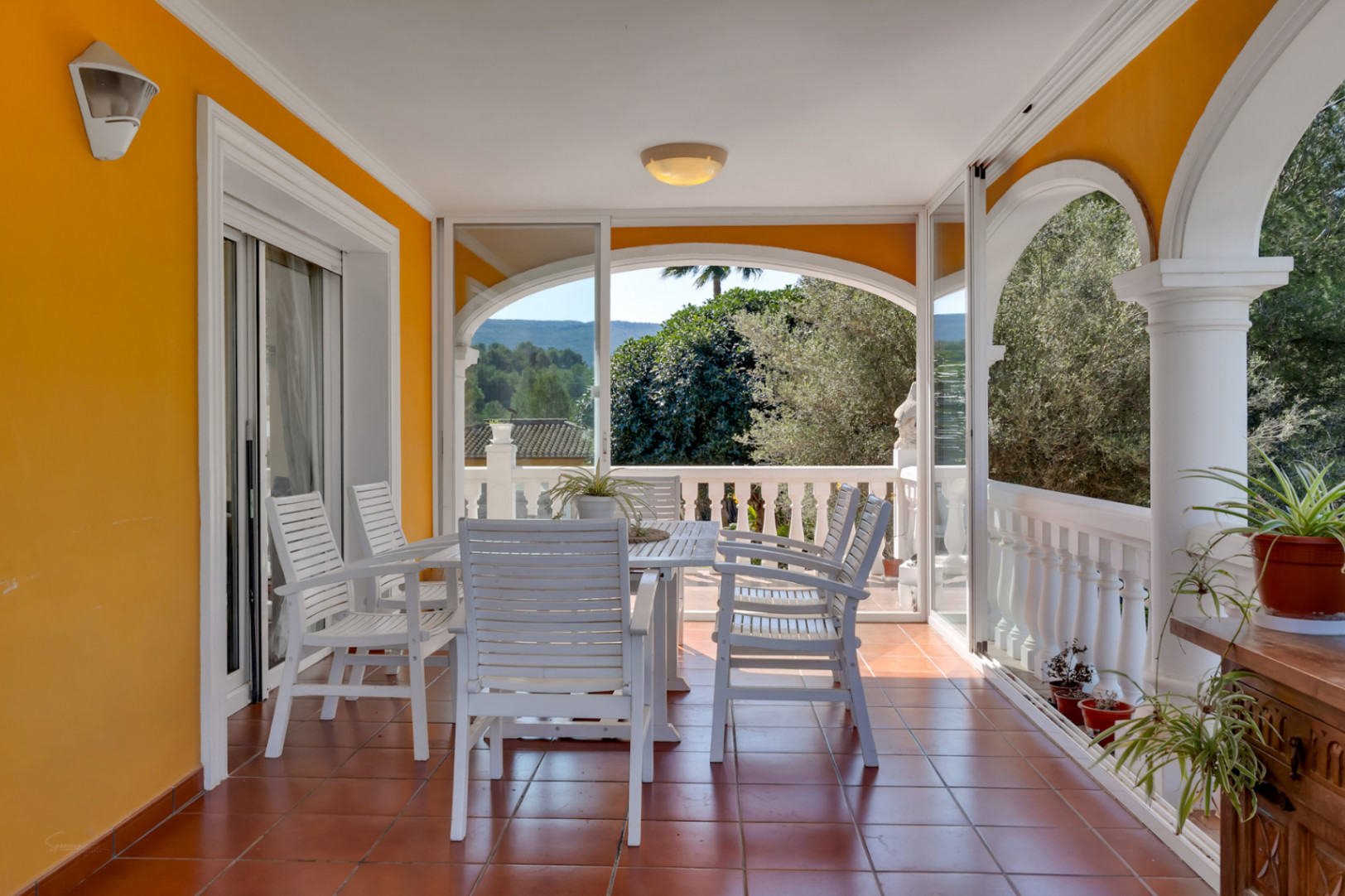 Terraza cubierta con zona de comedor, suelos de terracota y cerramientos acristalados que ofrecen vistas despejadas a la naturaleza y las montañas.