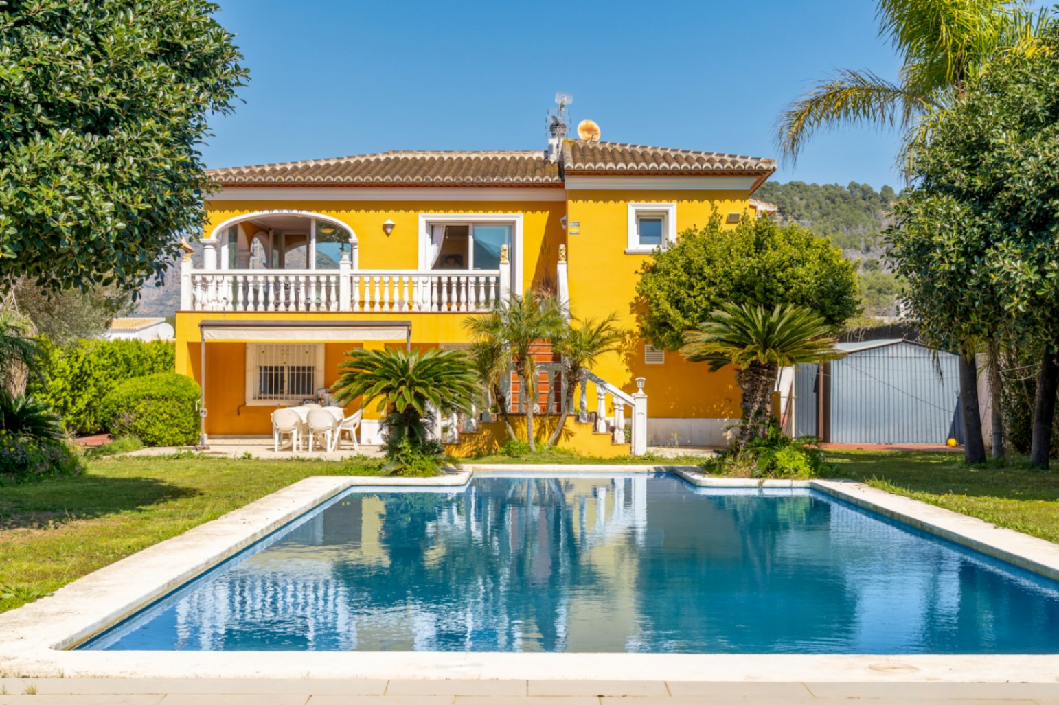Villa de fachada amarilla con amplia piscina rectangular, terraza superior con balaustrada blanca y jardín mediterráneo con palmeras y césped.
