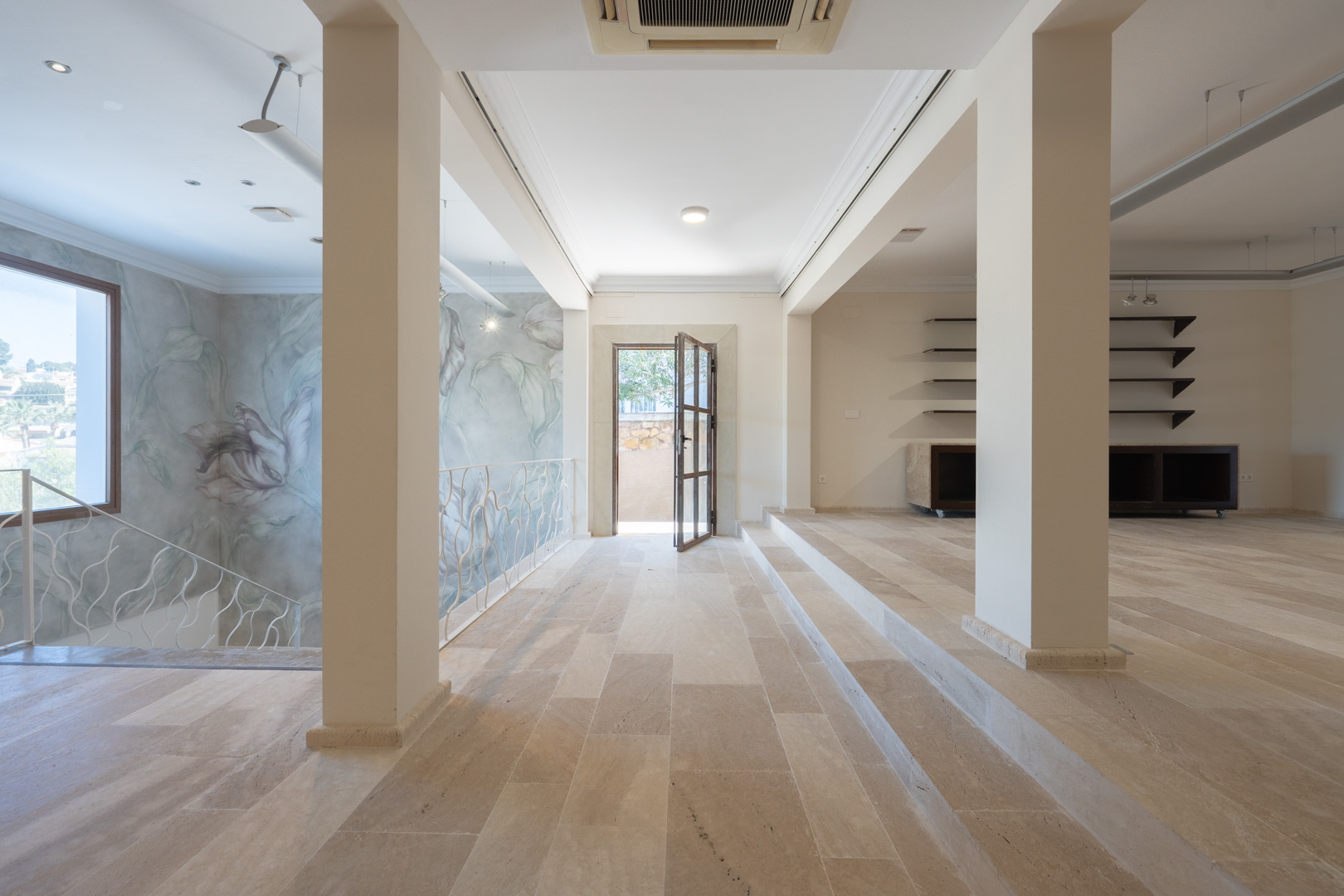 Amplio espacio diáfano en la primera planta con suelos de piedra natural, mural decorativo floral y barandilla de hierro forjado blanco junto a la escalera.