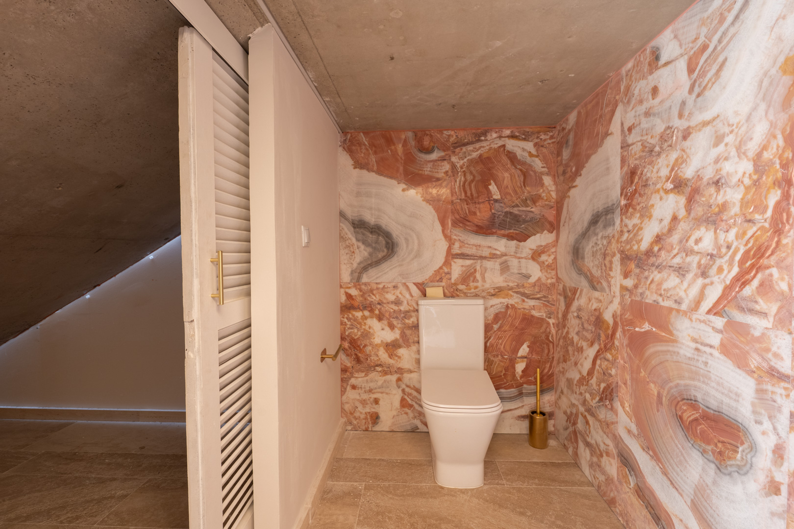Aseo con paredes de mármol veteado en tonos rosados, sanitario moderno y techo de hormigón visto. Incluye puerta corredera de lamas blancas y detalles en dorado.