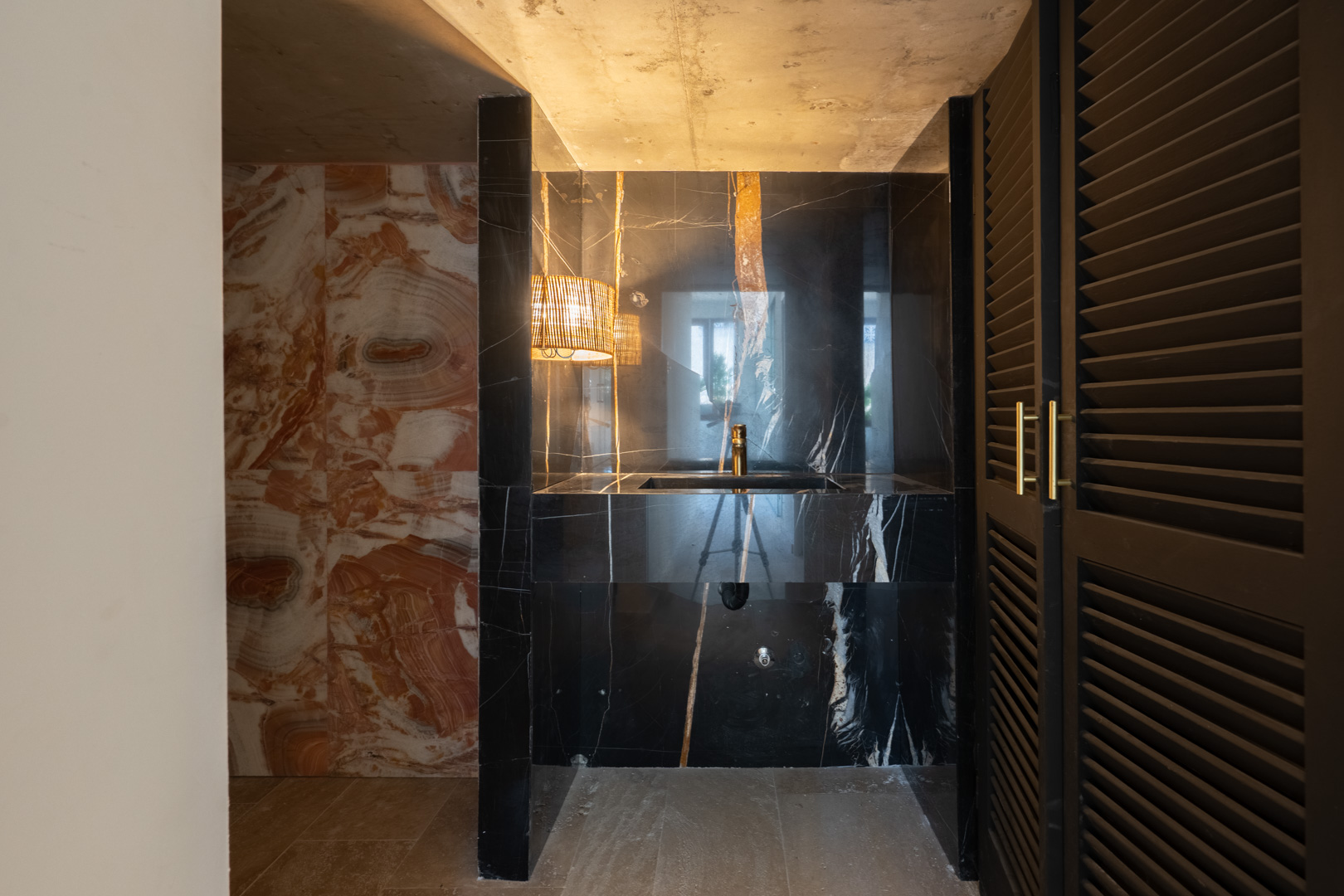 Aseo de diseño con lavabo integrado en mármol negro, grifería dorada y paredes de piedra natural veteada. Incluye iluminación ambiental y puertas de madera con lamas.
