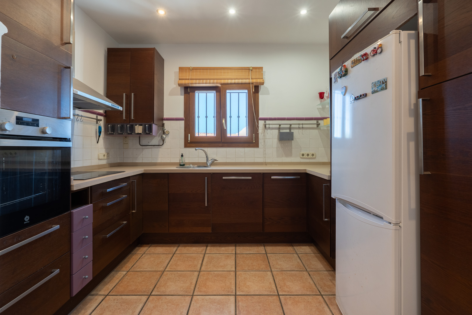 Cocina equipada con mobiliario de madera oscura, encimera en tonos crema y suelo de baldosas cerámicas. Dispone de electrodomésticos integrados y ventana con luz natural.