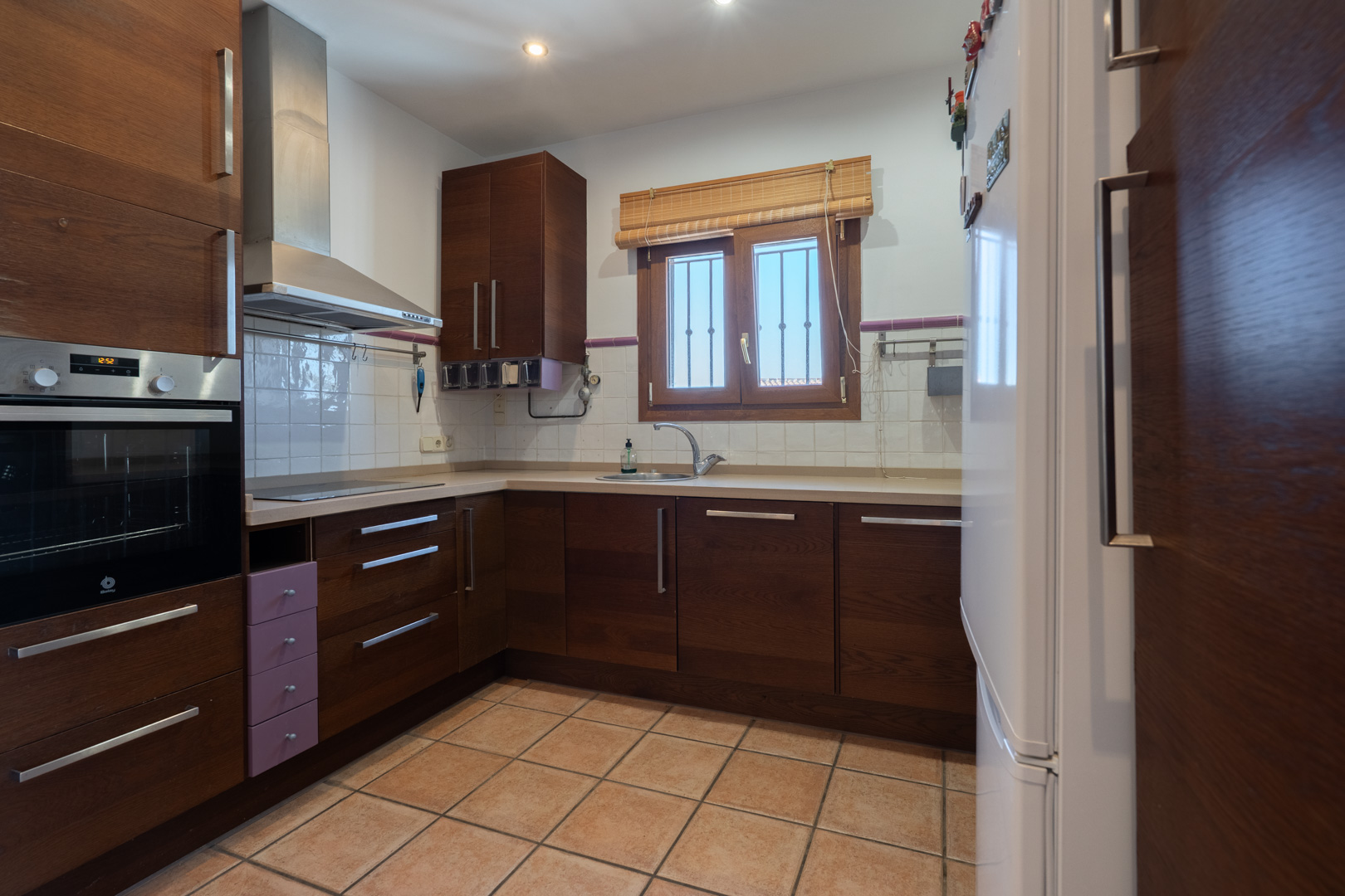 Cocina funcional con armarios de madera oscura, encimera clara y electrodomésticos integrados. Dispone de suelos cerámicos y ventana con luz natural sobre el fregadero.