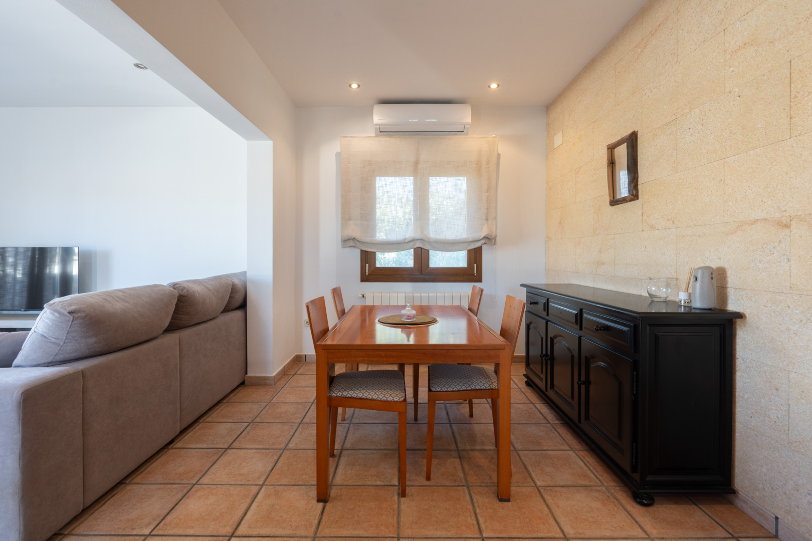 Comedor con mesa de madera, aparador negro y pared de piedra natural. Incluye suelos de terracota, aire acondicionado y acceso al salón contiguo.