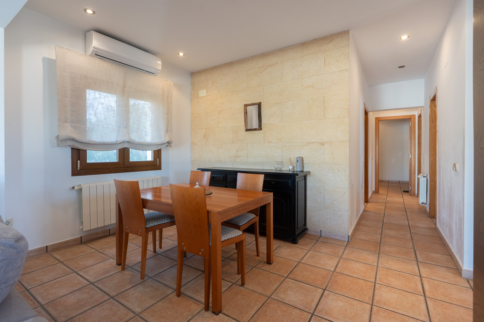 Comedor de la vivienda con pared de piedra natural, suelos de terracota, aire acondicionado y pasillo distribuidor hacia los dormitorios.