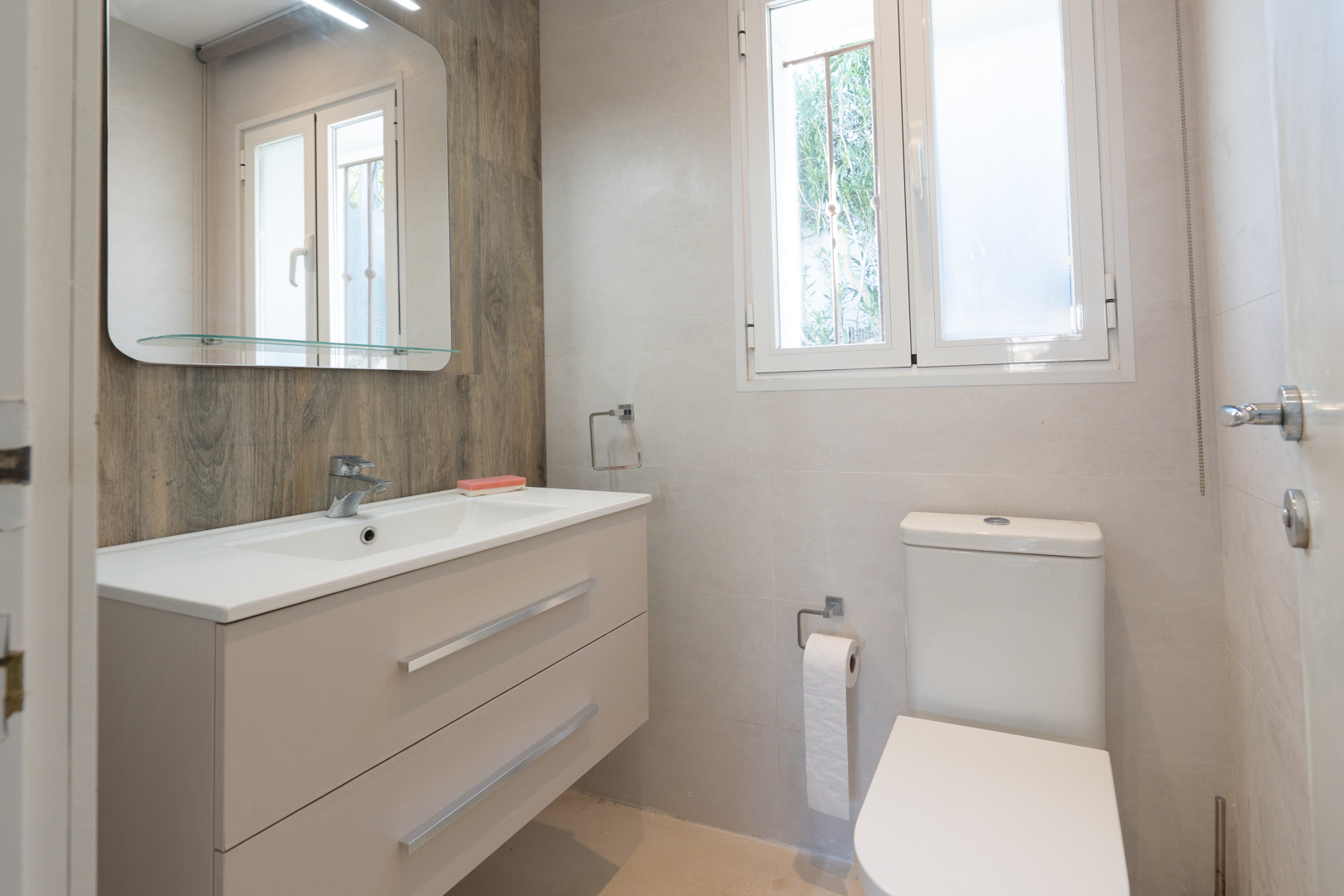 Cuarto de baño moderno con mueble de lavabo suspendido, inodoro blanco y pared de acento con efecto madera. Incluye ventana exterior para ventilación natural.