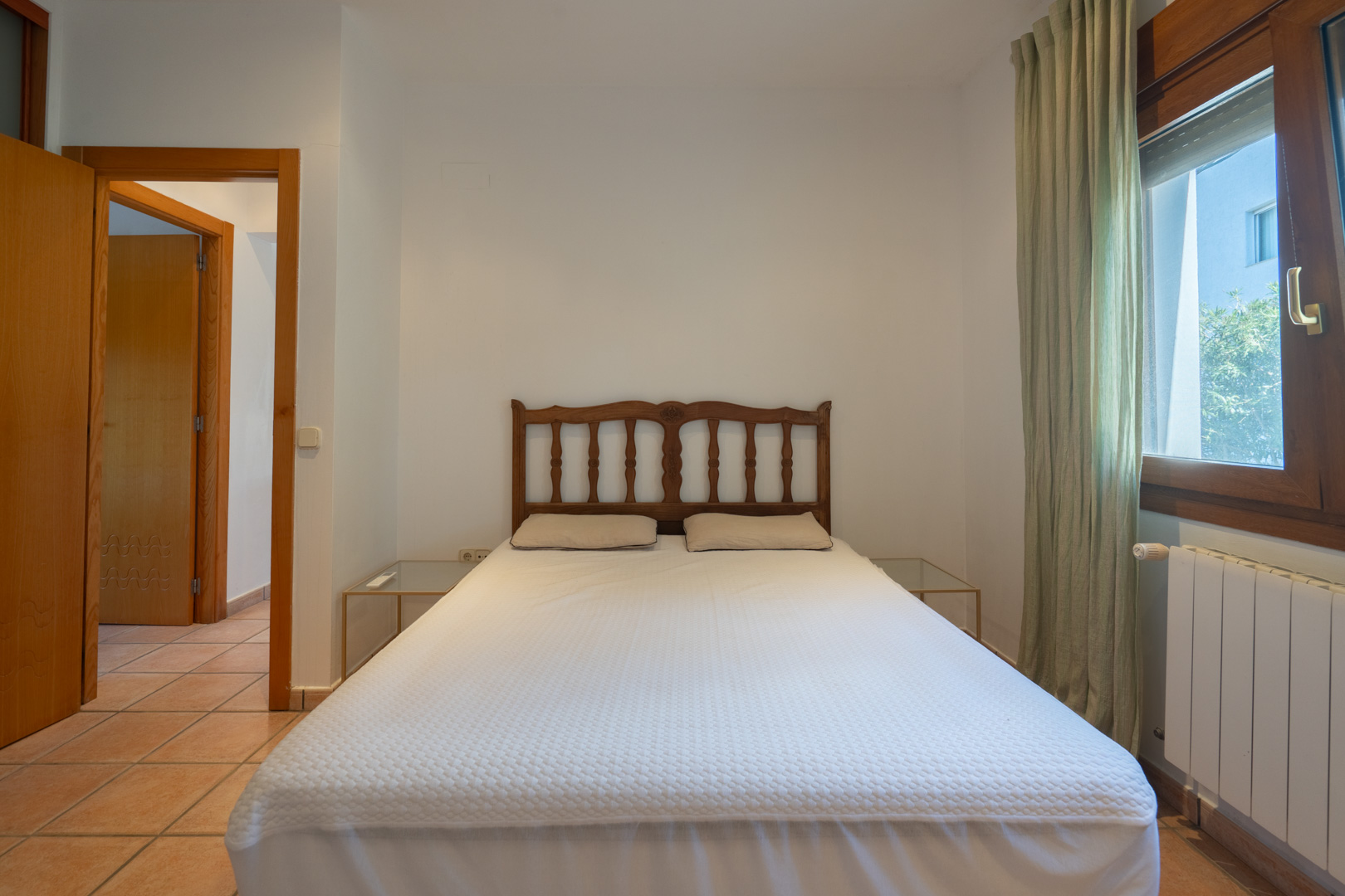 Dormitorio con suelos de baldosa cerámica, cabecero de madera tallada y ventana con marco de madera que aporta luz natural. Incluye radiador y mesas de noche de cristal.