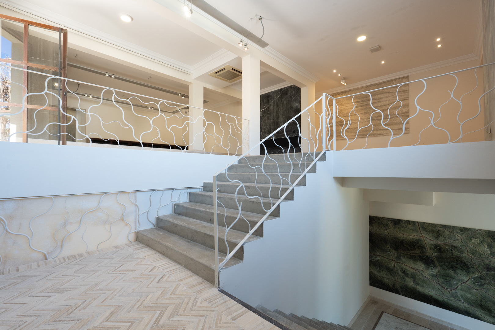 Escalera central con peldaños de piedra y barandilla de diseño orgánico, conectando los niveles comerciales con suelos de espiga y paneles de mármol.