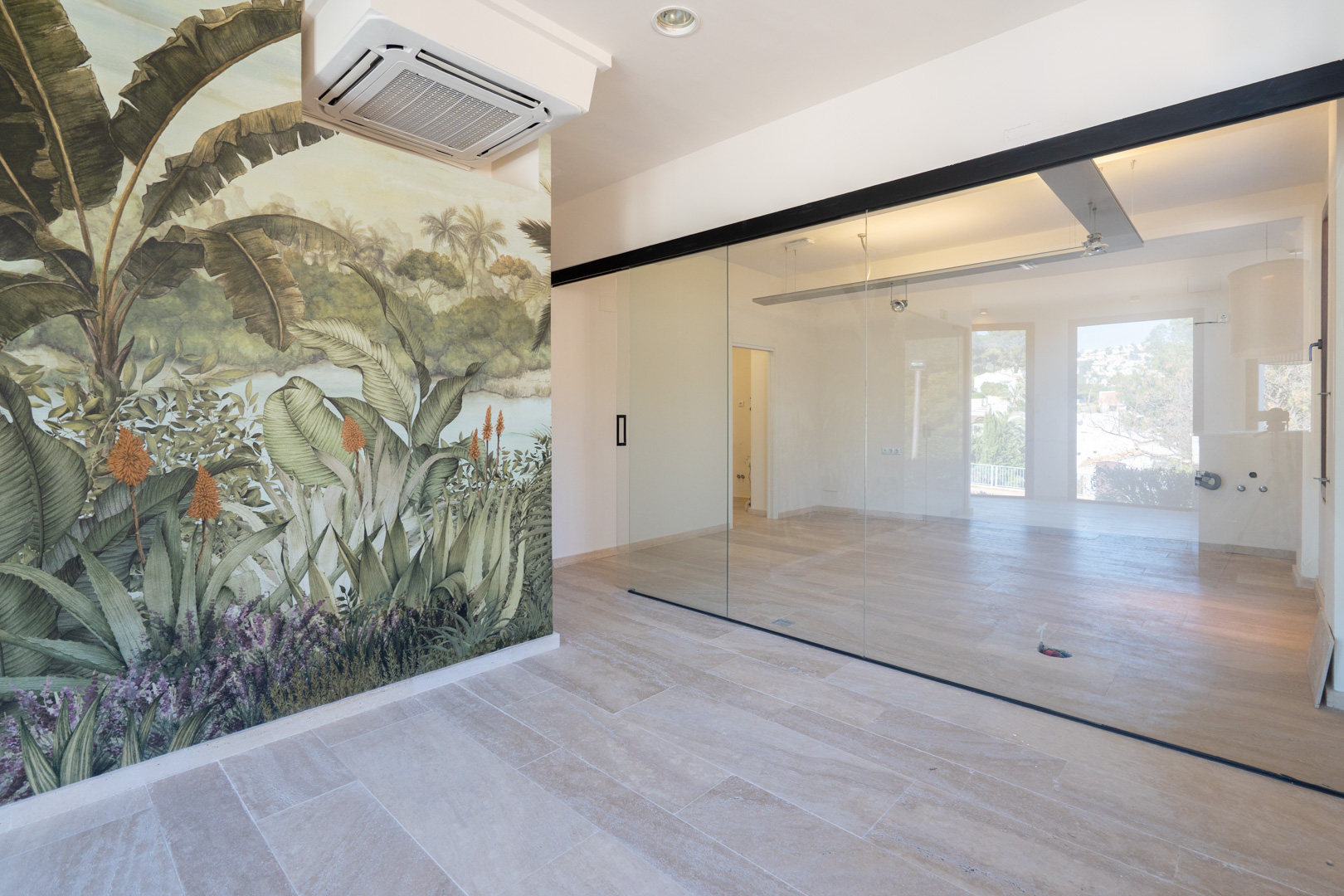 Espacio comercial diáfano con suelos de gres, mural decorativo tropical, cerramiento de cristal con perfilería negra y climatización por cassette en el techo.