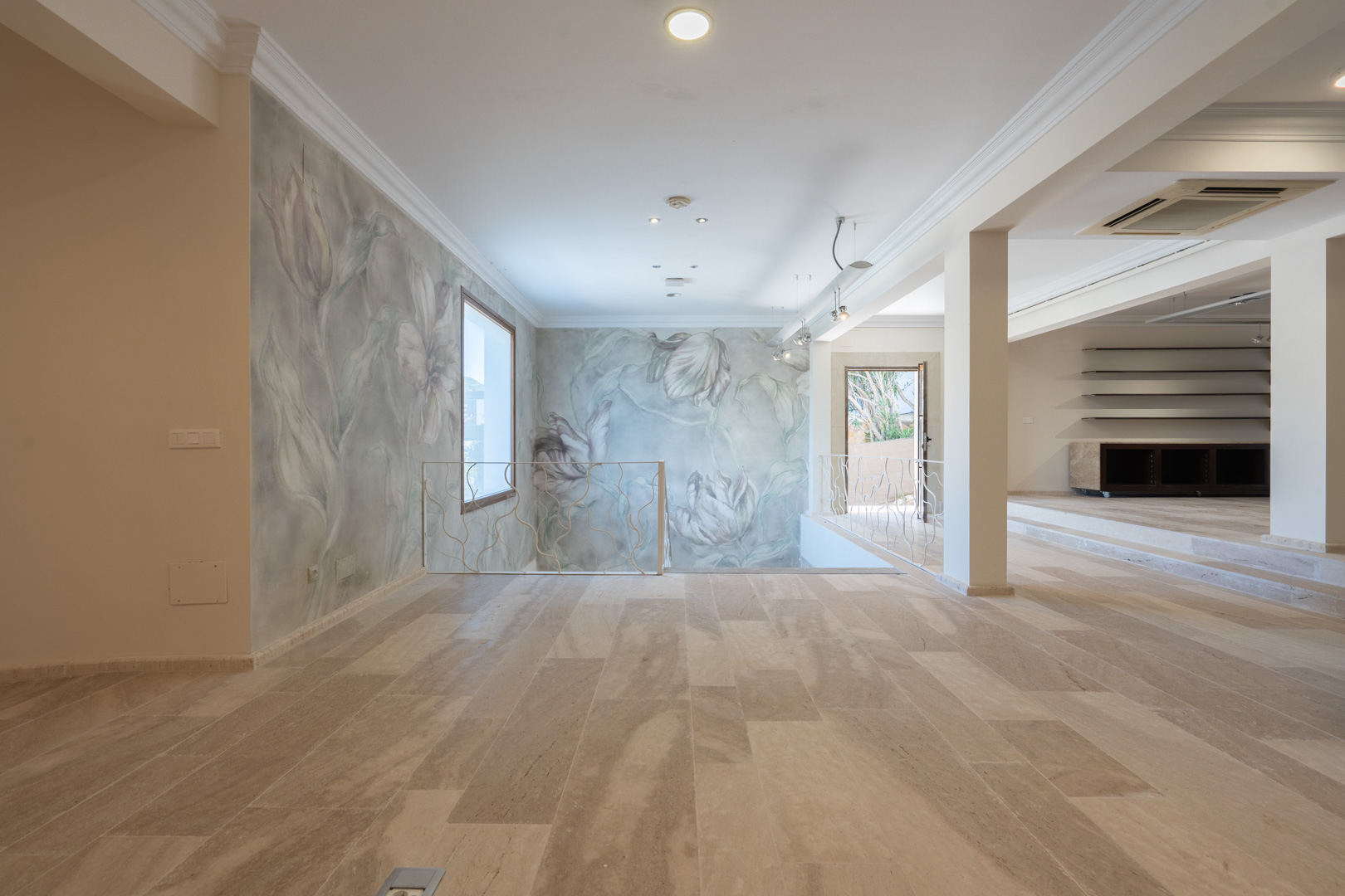 Espacio comercial diáfano con suelos de piedra natural, paredes decoradas con murales florales y una barandilla de hierro forjado blanco que delimita la escalera.