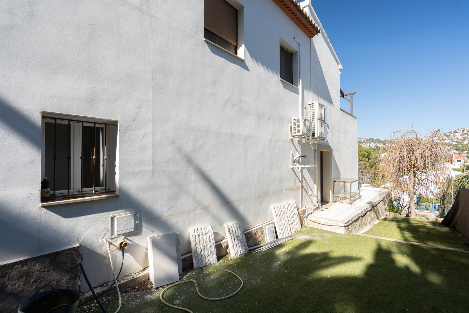 Fachada lateral blanca con base de piedra natural, unidades de aire acondicionado externas y zona de jardín con césped artificial y vistas despejadas al entorno.
