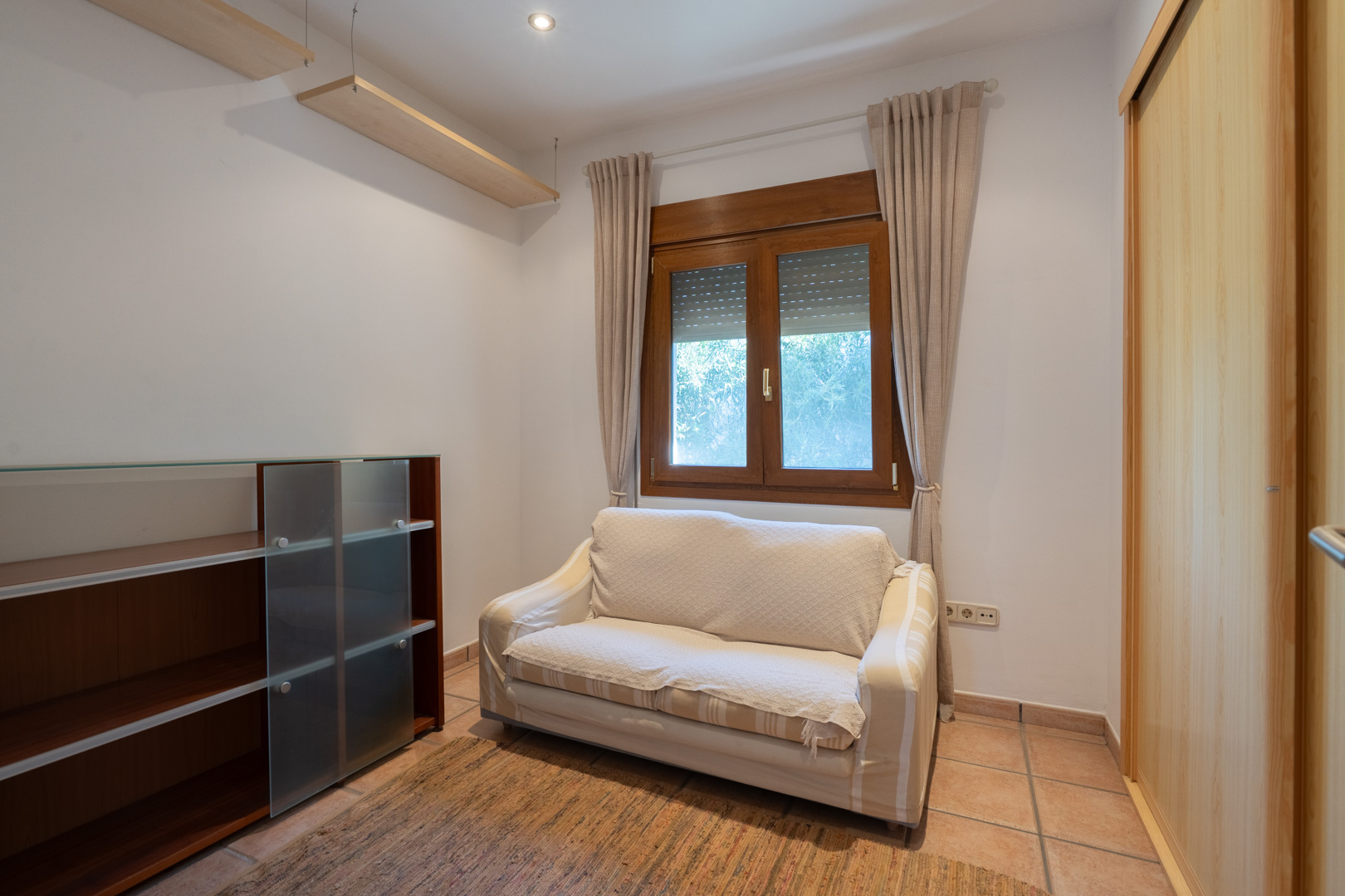 Habitación con suelos cerámicos, ventana de madera con persianas y armario empotrado. Dispone de estanterías de pared y un sofá pequeño en un ambiente funcional.