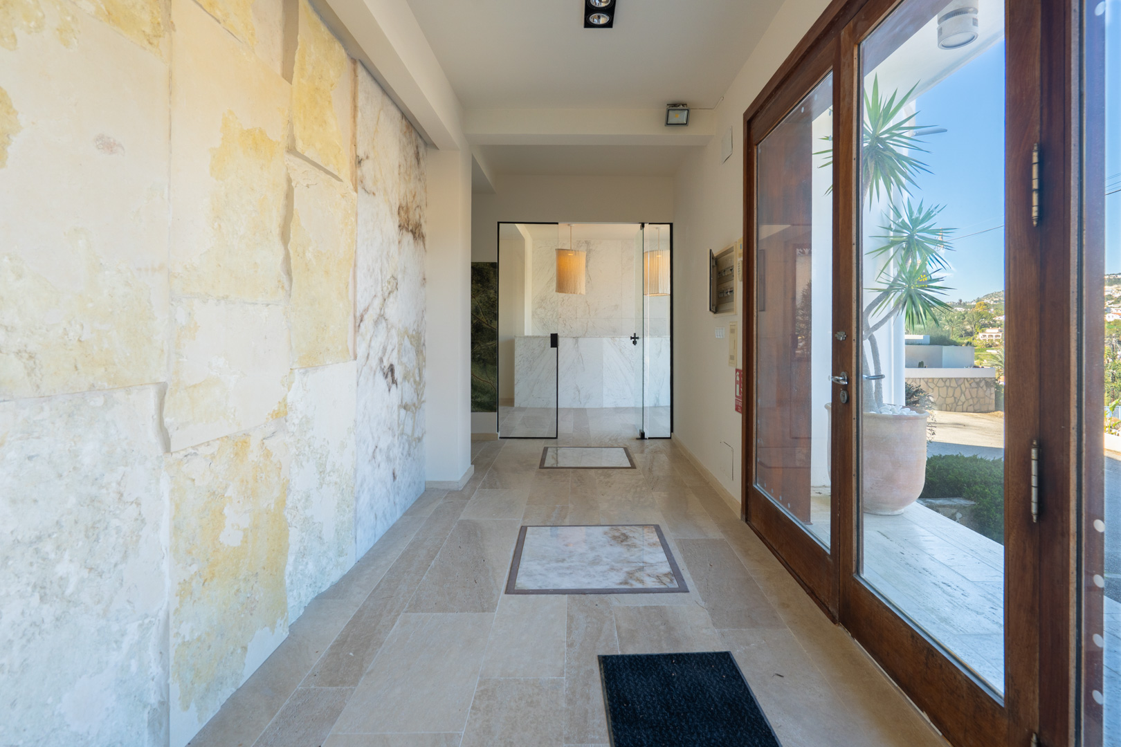 Hall de entrada con pared de piedra natural, suelos cerámicos con incrustaciones de mármol y grandes puertas acristaladas con marcos de madera.