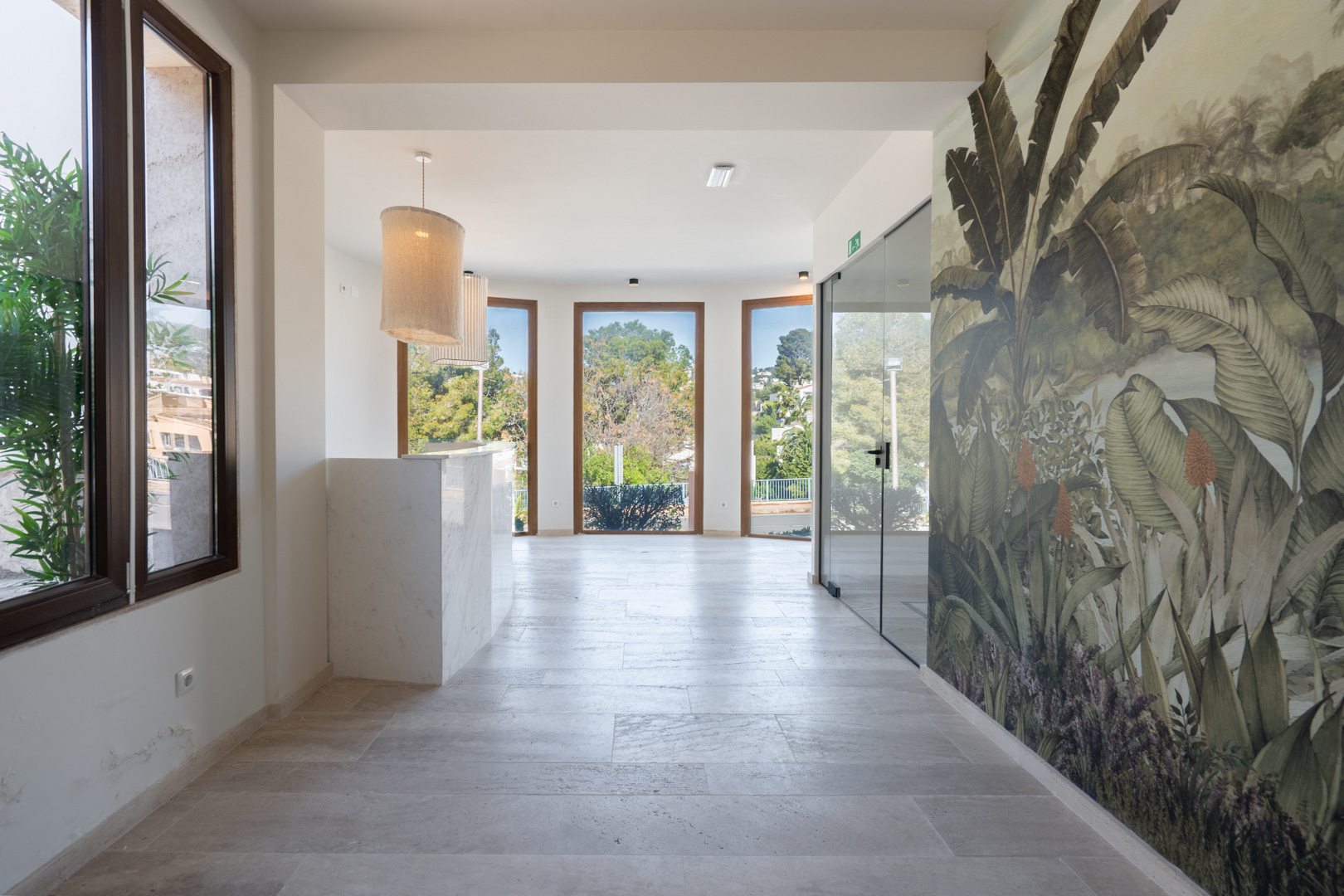 Hall de recepción con mostrador de mármol blanco, suelos de piedra natural y un mural tropical. Grandes ventanales con marcos de madera aportan abundante luz natural.