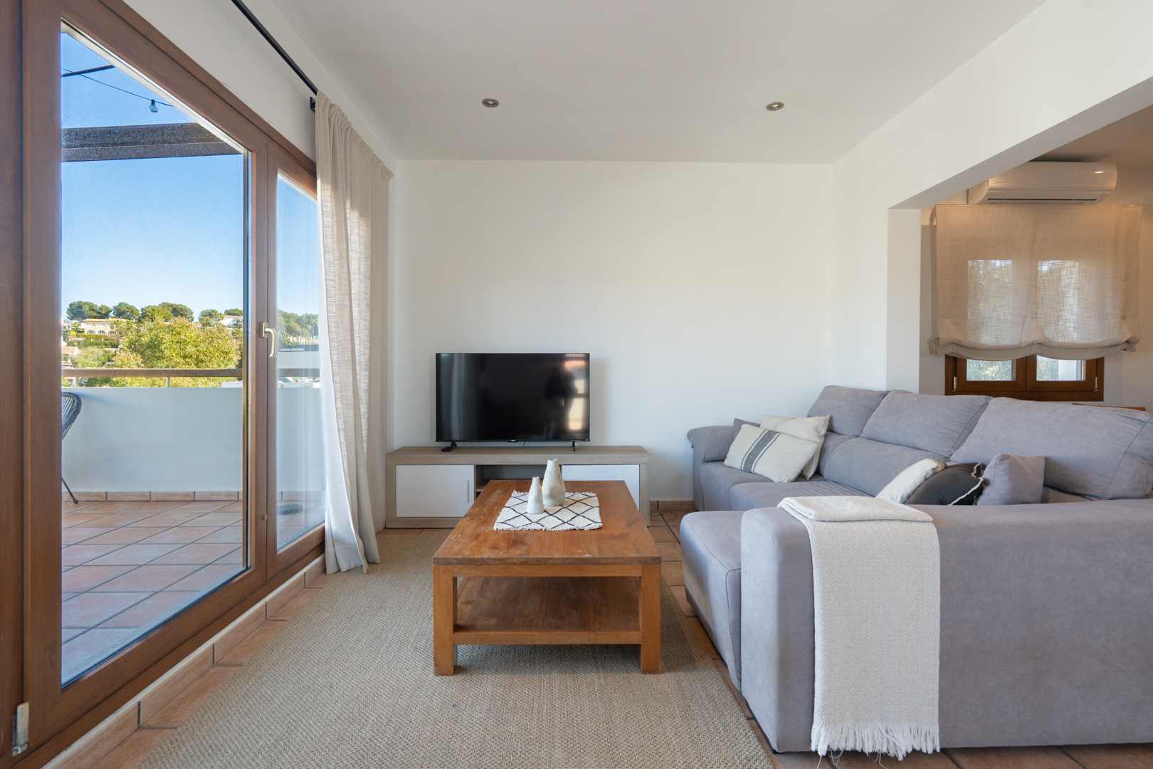 Salón luminoso con sofá gris, mesa de madera y acceso directo a la terraza mediante grandes ventanales con marco de madera y sistema de climatización.