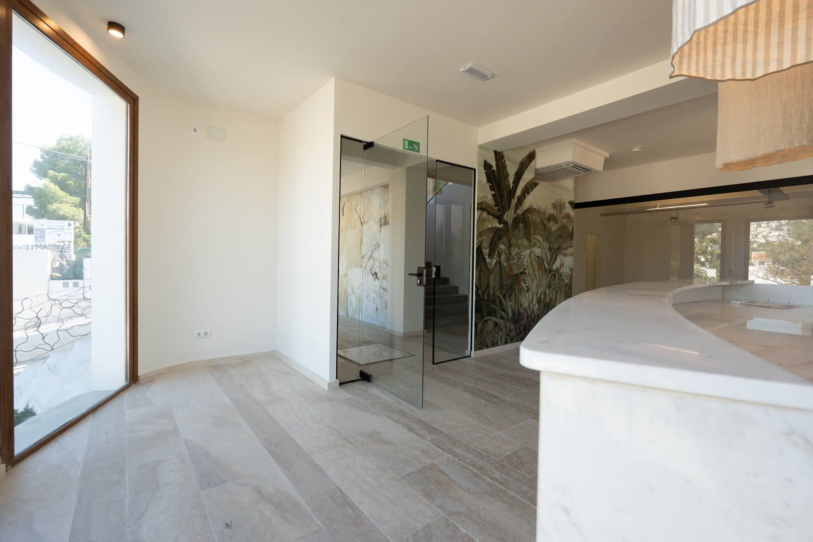 Zona de recepción comercial con mostrador de mármol blanco curvo, suelos de gres porcelánico, puertas de cristal y un mural decorativo de temática tropical.