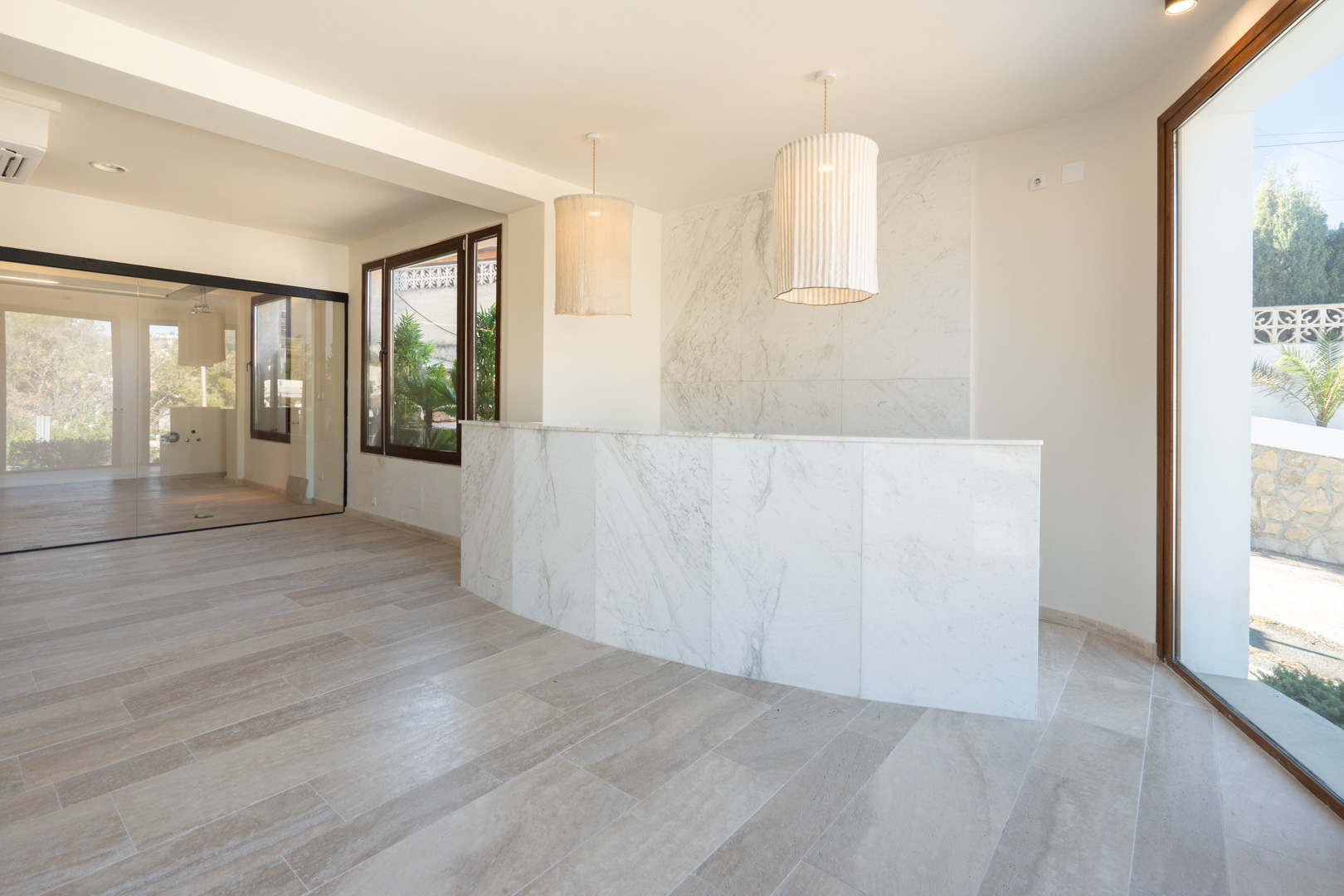 Zona de recepción con mostrador y pared de mármol, lámparas colgantes de tela y suelos de gres. Dispone de cerramientos de cristal y abundante luz natural.