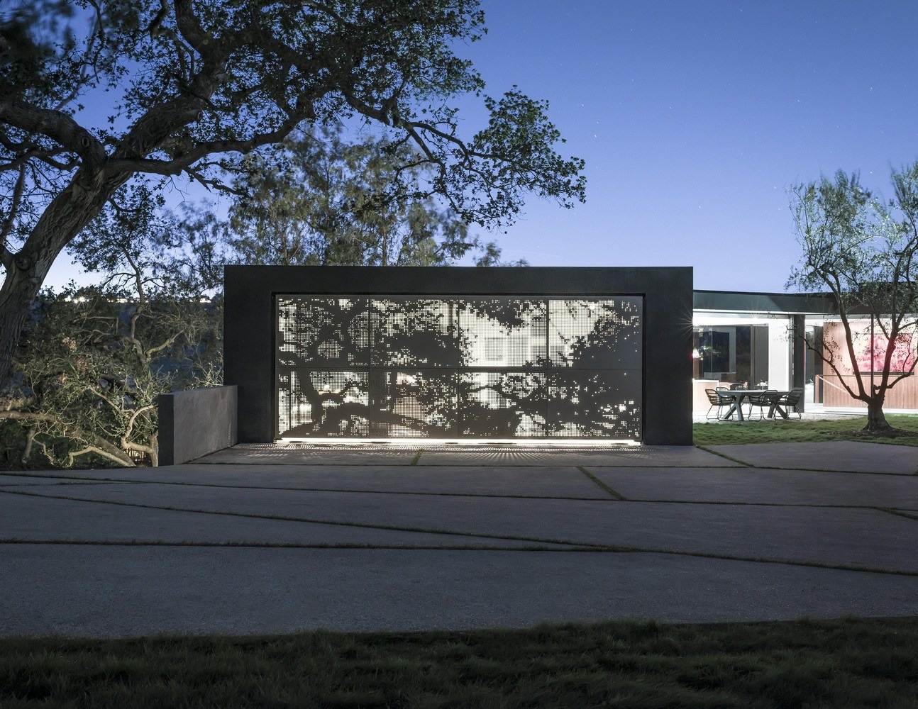 Fachada moderna con panel metálico perforado retroiluminado que proyecta siluetas de árboles, integrado en un entorno minimalista con pavimento de hormigón.