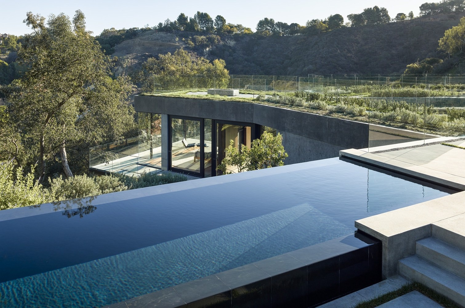 Piscina infinita de gresite oscuro integrada en una estructura de hormigón visto con cubiertas vegetales y amplios ventanales con vistas al entorno natural.