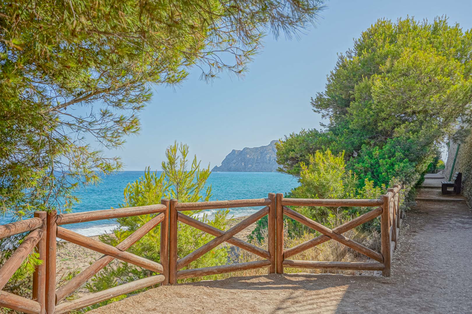 Sendero costero con valla de madera y vistas panorámicas al mar Mediterráneo y al Peñón de Ifach, rodeado de pinos autóctonos.