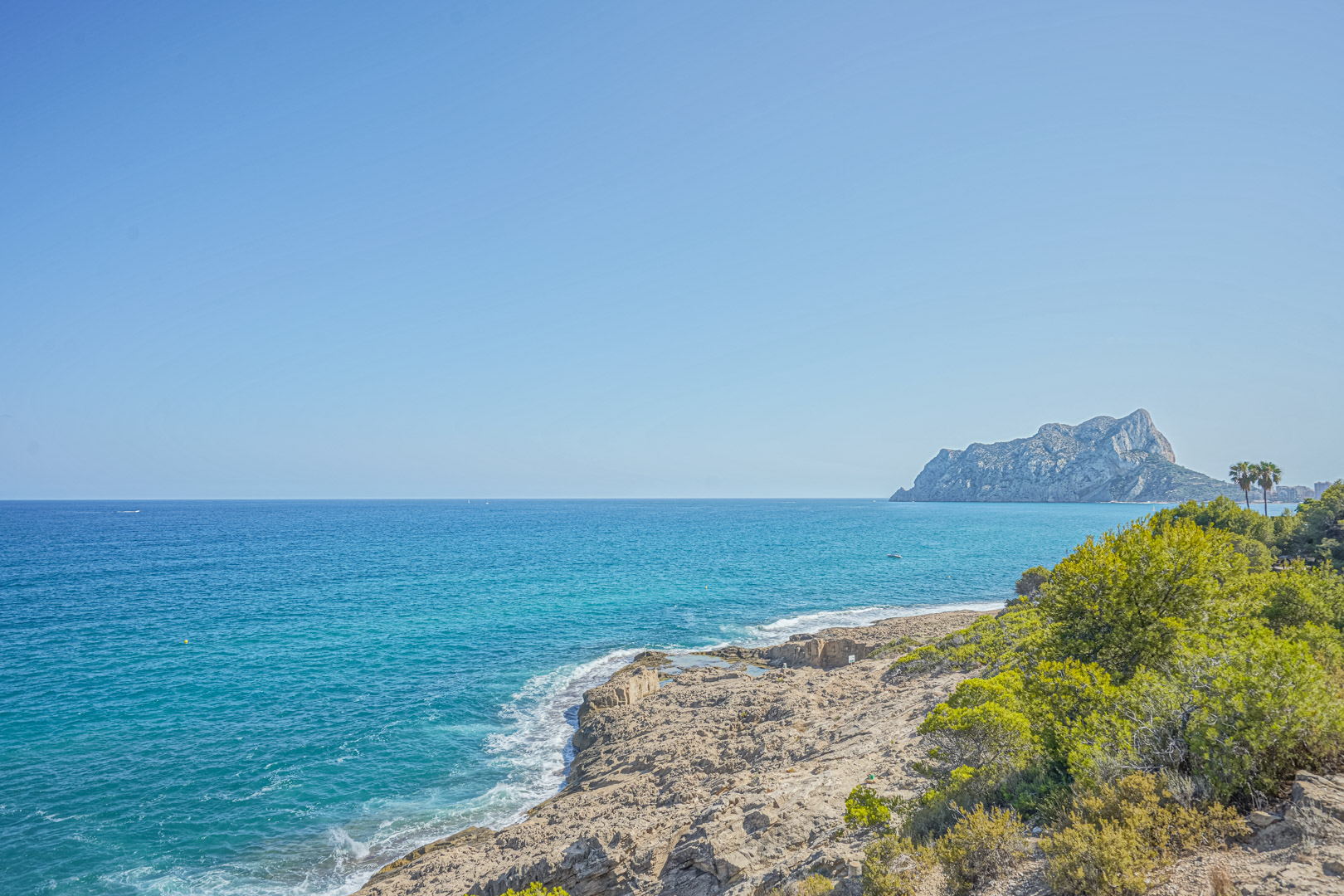 Vista panorámica de la costa de Benissa con el Peñón de Ifach al fondo, destacando las aguas cristalinas y la vegetación mediterránea junto a los acantilados rocosos.