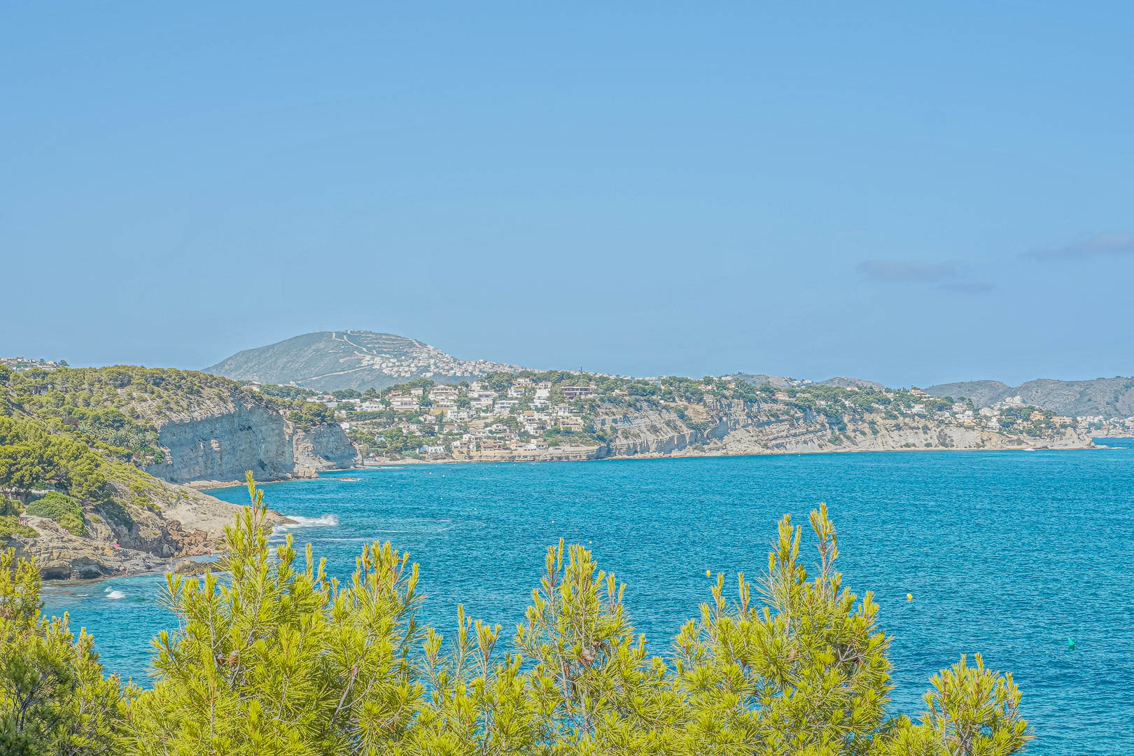 Vista panorámica de la costa de Benissa con pinos mediterráneos en primer plano y acantilados residenciales sobre las aguas turquesas del mar Mediterráneo.