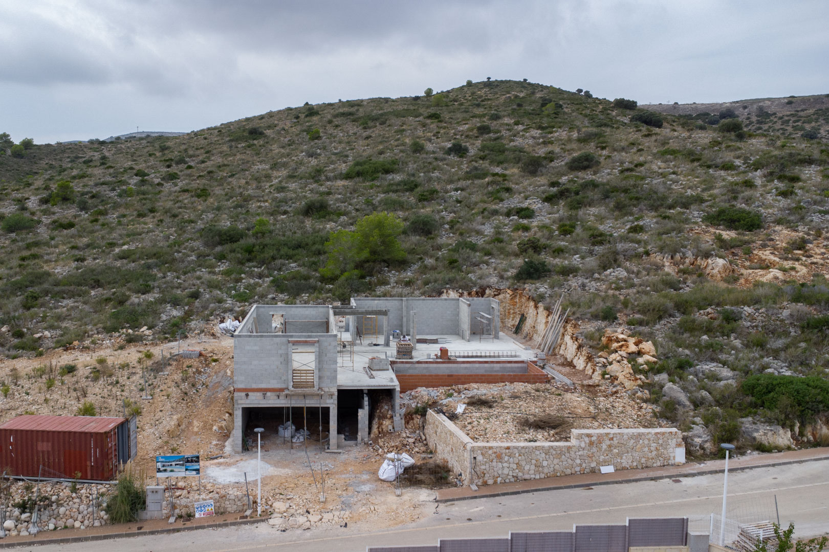 Fase de construcción de una villa multinivel en Pedreguer, con estructuras de bloques de hormigón, muro perimetral de piedra y ubicación en la ladera rocosa.