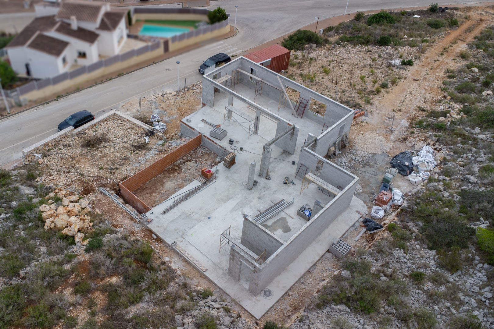 Vista aérea de los cimientos de hormigón y la estructura de muros de bloques de la villa en construcción, con materiales y andamios en la parcela.