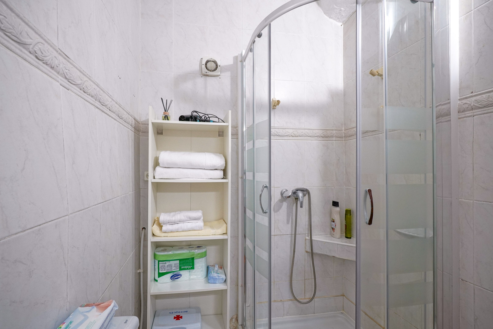 Baño funcional con plato de ducha curvo y mampara de cristal. Las paredes están revestidas con azulejos blancos y una cenefa decorativa, incluyendo una estantería de apoyo.