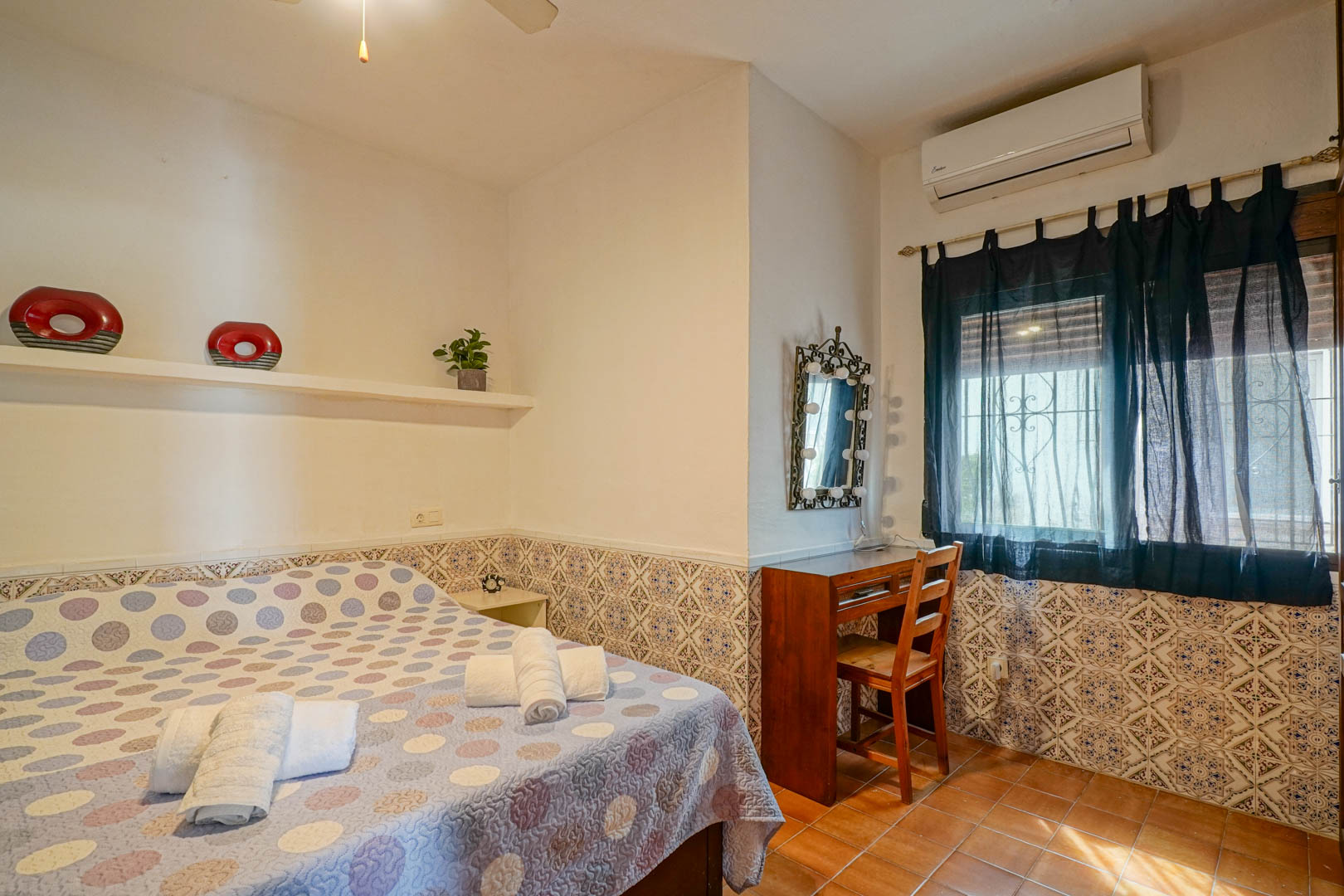 Dormitorio con zócalo de azulejos decorativos, escritorio de madera y espejo con iluminación. Incluye aire acondicionado y suelos de gres rústico.