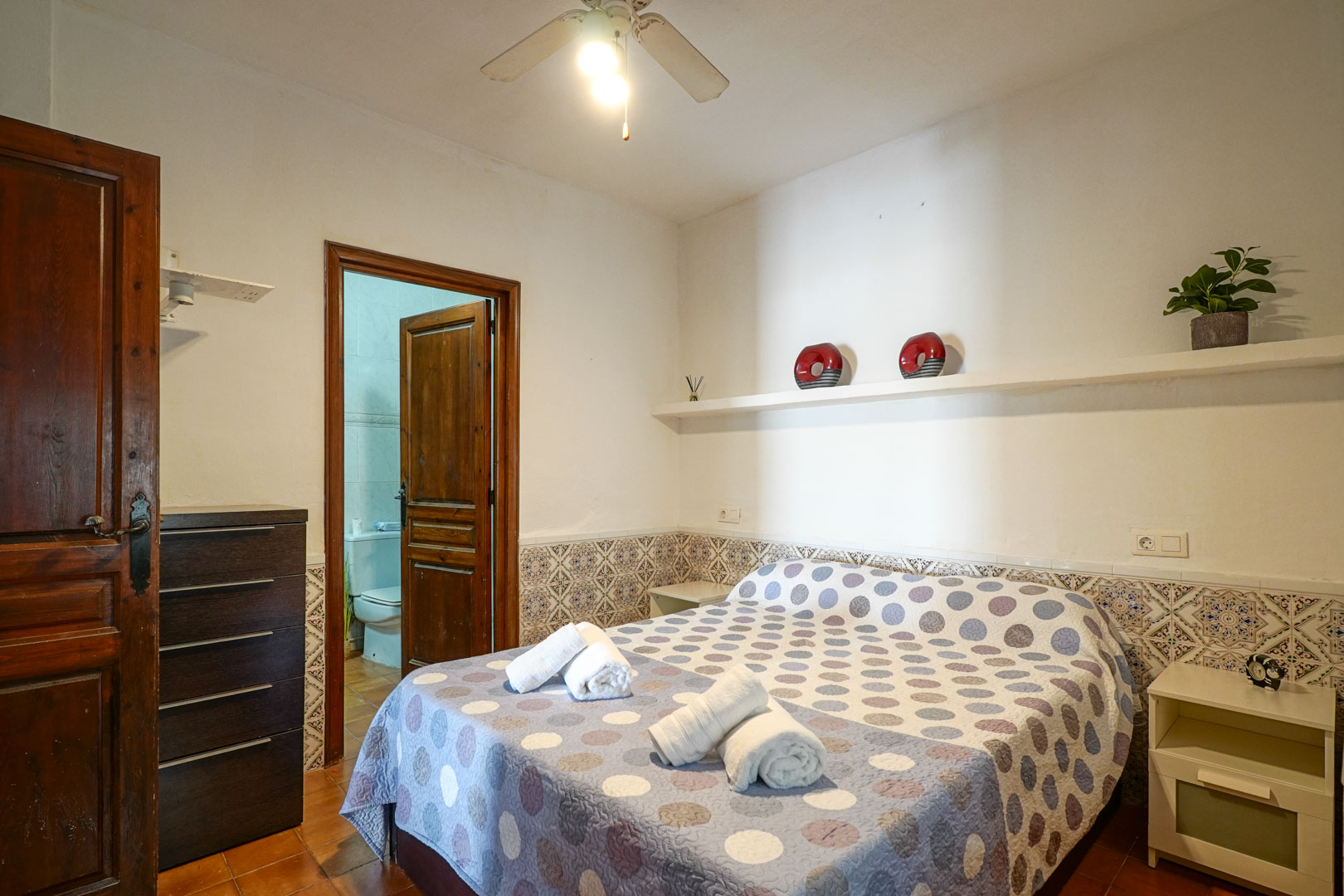Dormitorio principal tipo suite con zócalo de azulejos tradicionales, ventilador de techo y acceso directo al baño integrado a través de una puerta de madera.