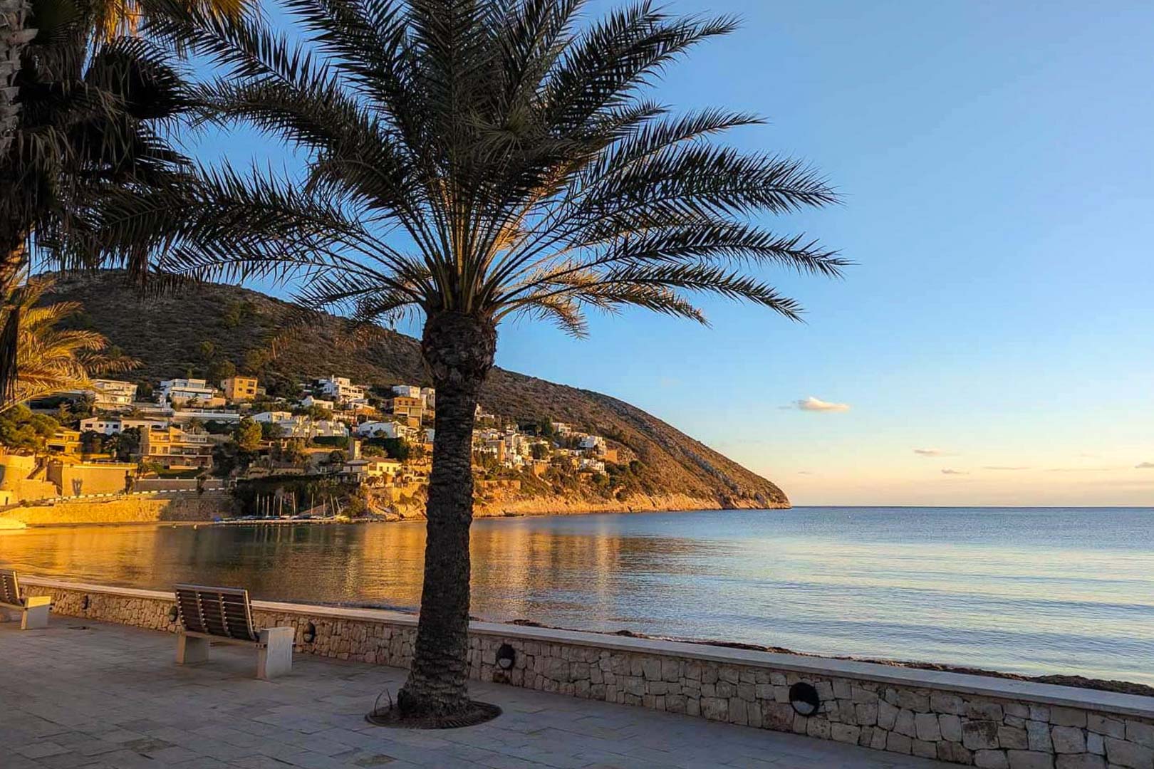 Paseo marítimo en Moraira con vistas a la bahía y colinas residenciales bajo la luz del atardecer. Incluye bancos de descanso, muros de piedra y vegetación mediterránea.