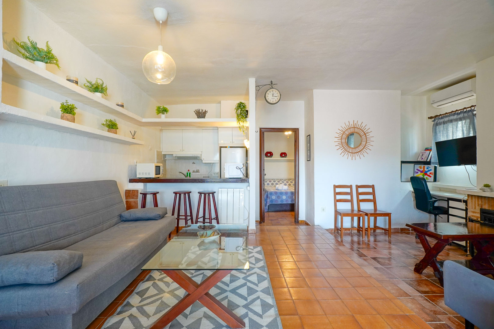 Salón comedor con cocina semiabierta, suelos de gres rústico y estanterías de obra. El espacio integra una zona de estar y un rincón de despacho junto al acceso al dormitorio.