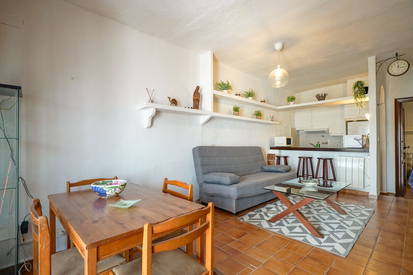 Salón comedor de concepto abierto con suelos de terracota, estanterías de obra blancas y cocina integrada con barra americana y taburetes de madera.
