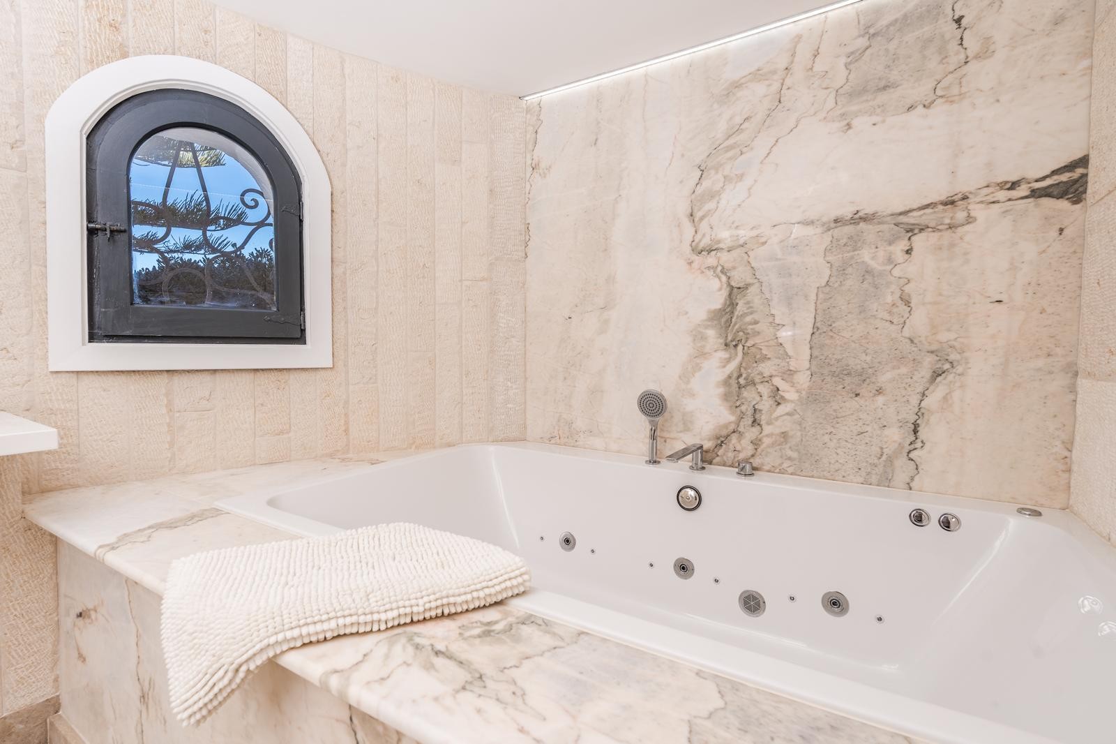 Bañera de hidromasaje encastrada en mármol veteado, situada junto a una ventana de arco tradicional y paredes de piedra natural con iluminación LED integrada.
