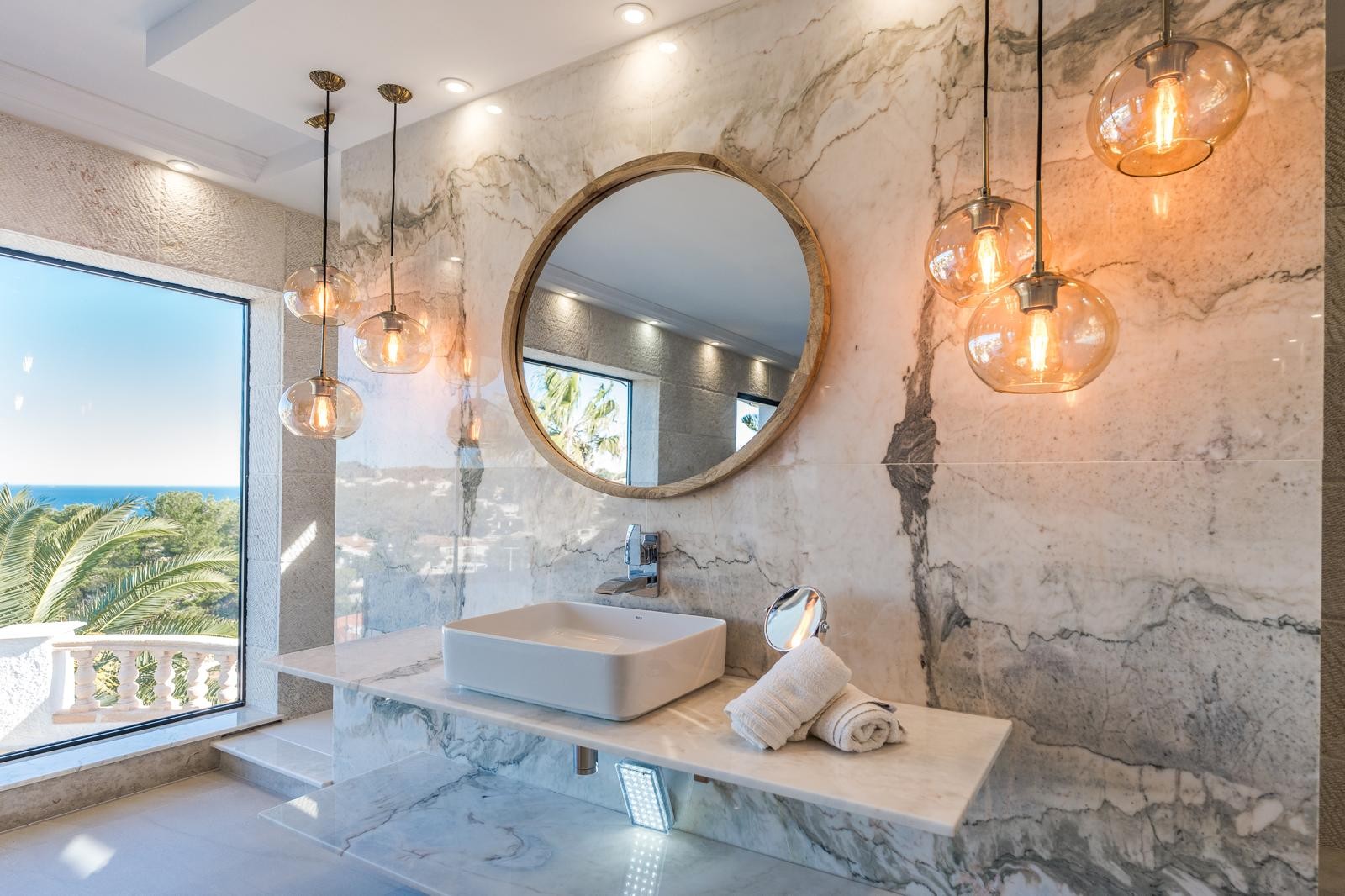 Baño de diseño con pared y encimera de mármol veteado, espejo circular con marco de madera y lámparas colgantes de cristal con vistas al mar.